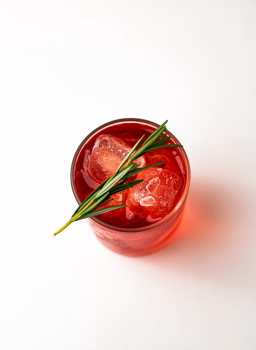 A red drink over large ice cubes in a short glass, garnished with a rosemary sprig