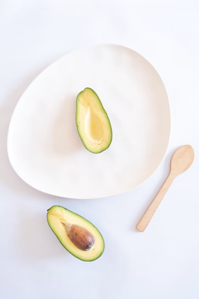 Two halves of an avocado on and beside a pale ceramic plate, with a wooden spoon