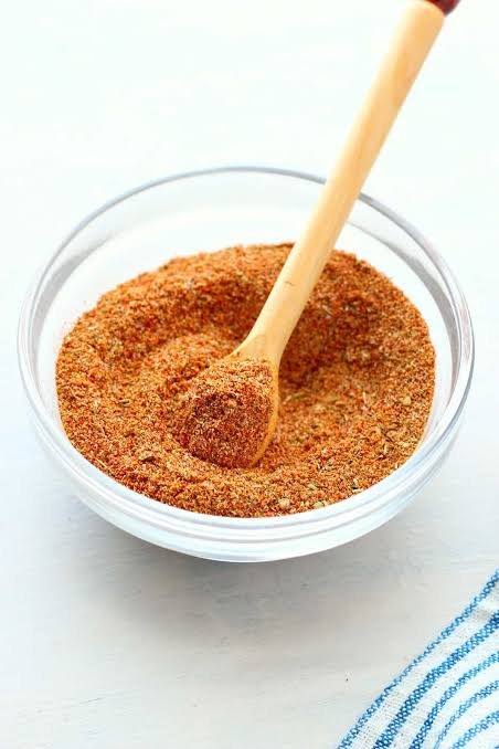 Reddish brown spice blend in a glass bowl with a small wooden spoon resting in it